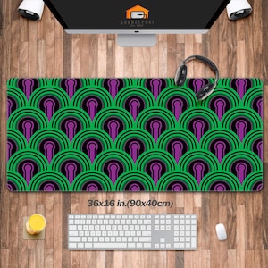 May include: A rectangular desk mat with a repeating geometric pattern of green, black, and purple semi-circles. The mat measures 36 x 16 inches. A keyboard, mouse, and headphones are on the mat. The mat is on a wooden desk.