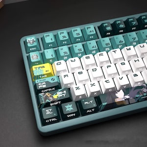 Puede incluir: Un teclado mecánico con marco verde azulado y una mezcla de teclas en verde azulado, blanco y verde oscuro. Las teclas muestran letras y gráficos estilizados, incluyendo una tecla tabulador amarilla y una ilustración de anime en la barra.
