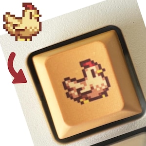 May include: A custom keyboard keycap featuring a pixelated chicken design in shades of tan, brown, and red. The keycap is a light orange color with a black border. A red arrow points to the chicken graphic.