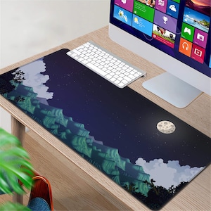 May include: A rectangular mouse pad featuring a pixelated landscape design with mountains, clouds, and a moon against a dark blue sky. A white keyboard and computer monitor are visible on a wooden desk.