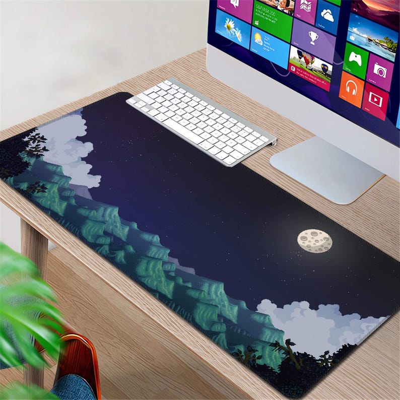 May include: A rectangular mouse pad featuring a pixelated landscape design with mountains, clouds, and a moon against a dark blue sky. A white keyboard and computer monitor are visible on a wooden desk.