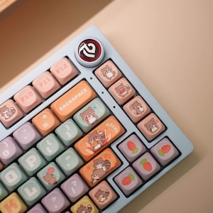 May include: A light blue mechanical keyboard with custom keycaps. The keycaps feature a variety of colors and illustrations of raccoons and carrots. The keyboard also has the text "BACKSPACE" and "enter" on the keys.