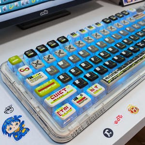 May include: A custom mechanical keyboard with transparent keycaps and blue LED backlighting. The keycaps feature various icons and text, including "ESC", "TAB", and "WIN". The keyboard sits on a white surface with stickers.