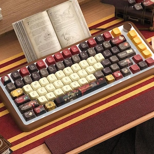 Pode incluir: Um teclado mecânico personalizado com uma moldura castanha e teclas brancas, castanhas e vermelhas. O teclado está colocado numa superfície listrada a vermelho e dourado, com um livro aberto atrás. Algumas teclas têm texto e gráficos.