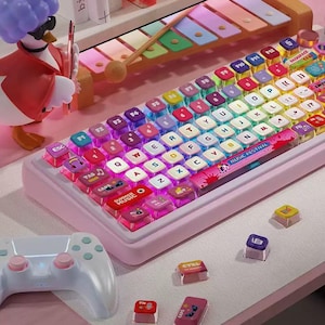 Puede incluir: Un teclado mecánico rosa con teclas multicolores. Las teclas están etiquetadas con letras, números y símbolos. El teclado tiene una base rosa y un diseño "MUSIC FESTIVAL". También se ve un mando de juego blanco y teclas adicionales.