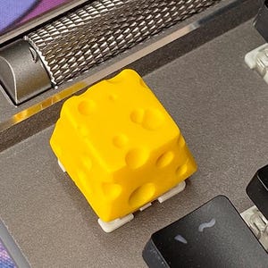 May include: A bright yellow, cheese-shaped keycap with holes, on a grey mechanical keyboard. The keycap is a vibrant yellow and has a textured surface. Other black keys are visible.