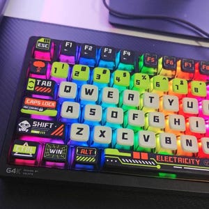 May include: A black G4K mechanical keyboard with vibrant, backlit keys. The keys are illuminated in various colours, including pink, blue, green, and yellow. The keyboard features the word "ELECTRICITY".