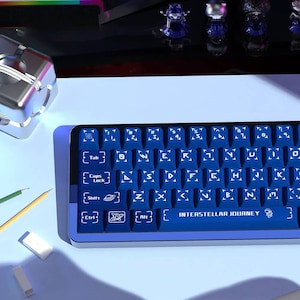 May include: A blue and purple mechanical keyboard with white keycap legends. The keyboard has a compact layout and features the text "INTERSTELLAR JOURNEY" on the spacebar. Other items in the image include pencils, erasers, and decorative figurines.