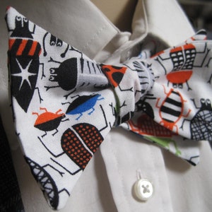May include: A white bow tie with a black and orange bug pattern. The bow tie is made of fabric and has a white background with black and orange bugs on it.