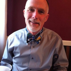 May include: A man with a white beard is wearing a light blue button-down shirt with a colorful bow tie. He is smiling at the camera.