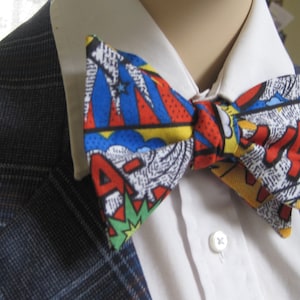 May include: A blue, red, yellow, and white bow tie with a comic book pattern. The bow tie is tied around a white collared shirt and a blue plaid jacket.