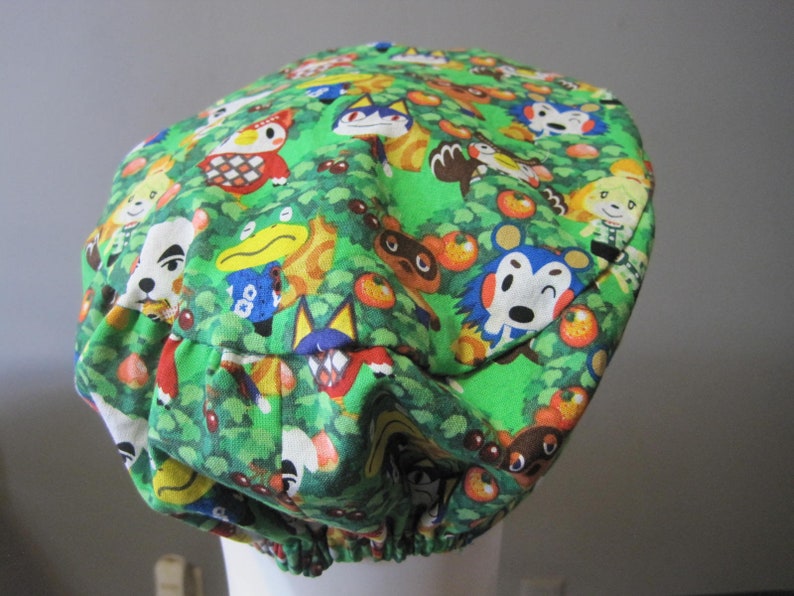 May include: A green fabric hat with a colorful pattern of animal characters from the Animal Crossing video game series. The hat has a gathered elastic band around the bottom.