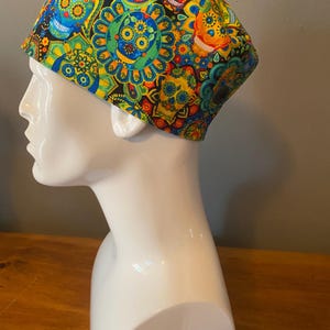 May include: A colorful skull cap featuring a vibrant pattern of smiling skulls, paisley shapes, and floral designs in shades of yellow, blue, orange, and green. The cap is set against a neutral background.