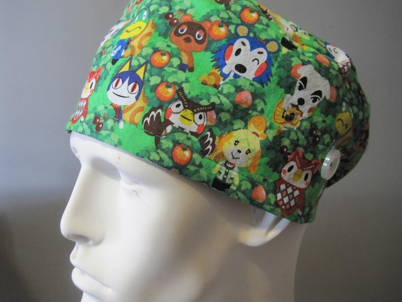 May include: Green scrub hat with a colorful print of animal characters from the video game Animal Crossing. The hat has a button on the side.