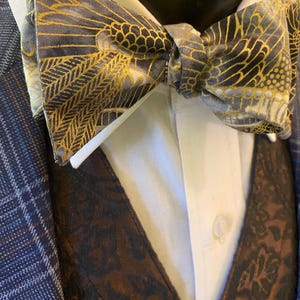 May include: A patterned bow tie with gold and black accents, paired with a white dress shirt and a brown vest. The bow tie features a detailed design, adding a touch of elegance to the ensemble. The vest has a dark floral pattern.