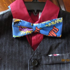 May include: A black and white striped vest with a red button and a pocket. A red shirt with a blue bow tie with a comic book print. The bow tie has the words "WOW", "POW", and "BAM" printed on it. The date "08/10/2014" is printed on the vest.