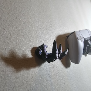 May include: A white video game controller mounted on a wall with a 3D printed black hand holding it.
