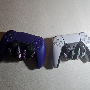 May include: Two video game controllers, one purple and one white, with a 3D printed claw-like grip accessory attached to each.