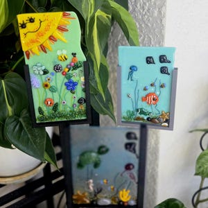 May include: Three decorative glass art pieces with black frames. One features a yellow sun and colorful flowers, another depicts an underwater scene with fish, and the third showcases a sea turtle. The art is set against a backdrop of green plants.