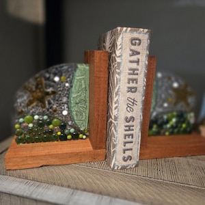 May include: Wooden bookends with a decorative design. The bookends feature a central wooden support and two decorative sides with glass and bead embellishments. The book on the right has the text "GATHER the SHELLS".
