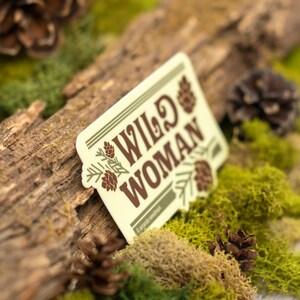 May include: A rectangular sticker with the words "WILD WOMAN" in brown lettering, accented with pine cone illustrations. The sticker is set against a backdrop of moss, a wooden log, and pine cones, creating a nature-inspired theme.