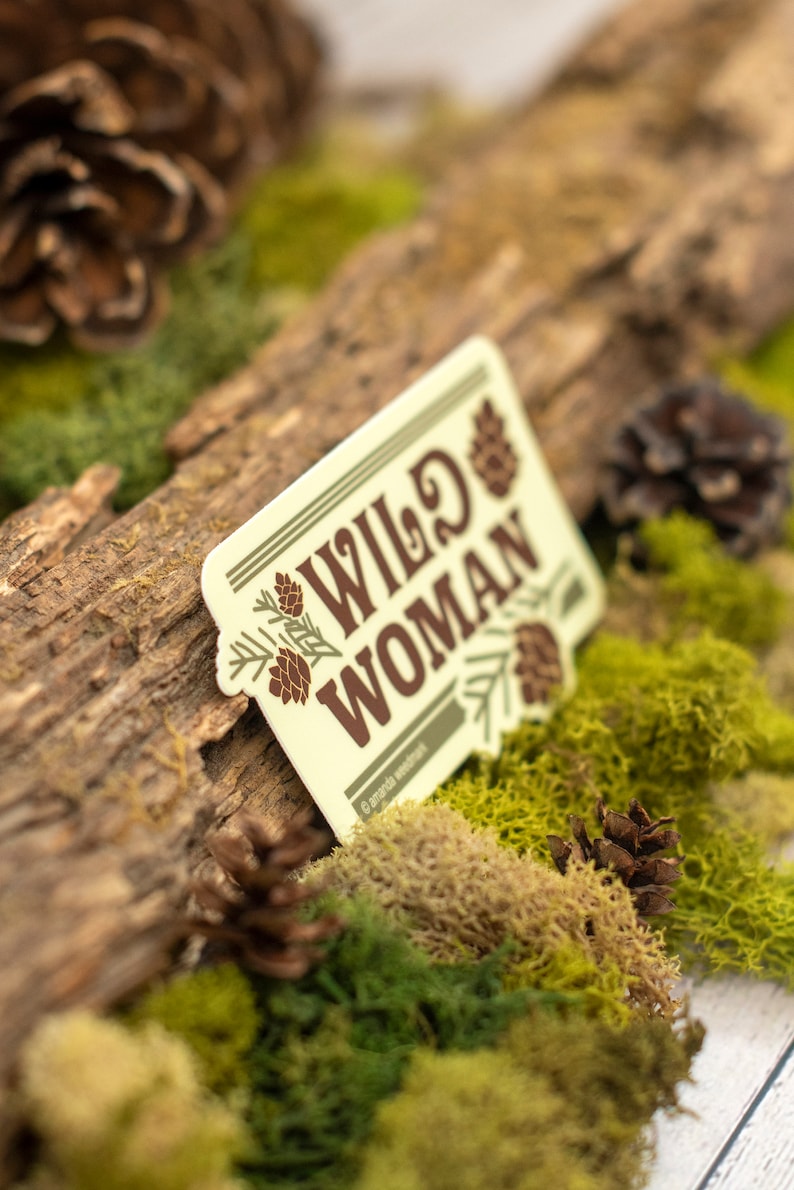 May include: A rectangular sticker with the words "WILD WOMAN" in brown lettering, accented with pine cone illustrations. The sticker is set against a backdrop of moss, a wooden log, and pine cones, creating a nature-inspired theme.