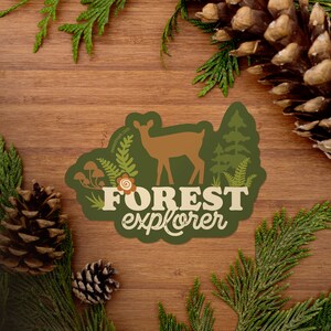 May include: A green sticker with a brown deer silhouette, green trees, and mushrooms. The text "FOREST explorer" is in white with a brown outline.