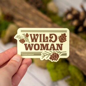 May include: A rectangular sticker with the words "WILD WOMAN" in brown, stylized font. The sticker has a light yellow background with brown pinecone and branch illustrations. The sticker is held in a hand.