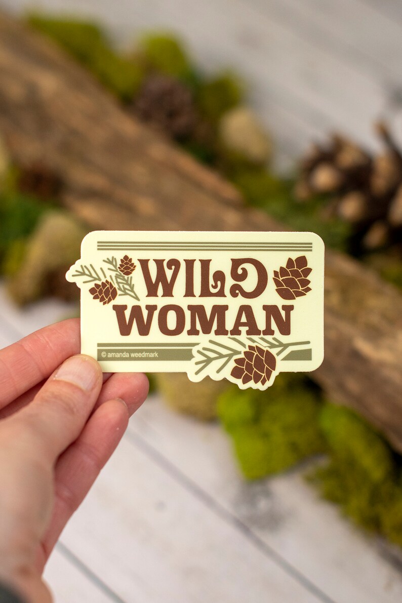 May include: A rectangular sticker with the words "WILD WOMAN" in brown, stylized font. The sticker has a light yellow background with brown pinecone and branch illustrations. The sticker is held in a hand.