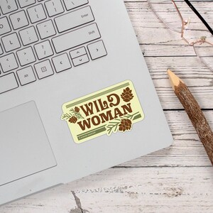 May include: A rectangular sticker on a laptop with the words "WILD WOMAN" in brown, surrounded by pine cones and branches. The sticker has a light yellow background and a green border. A pencil and glasses are also visible.
