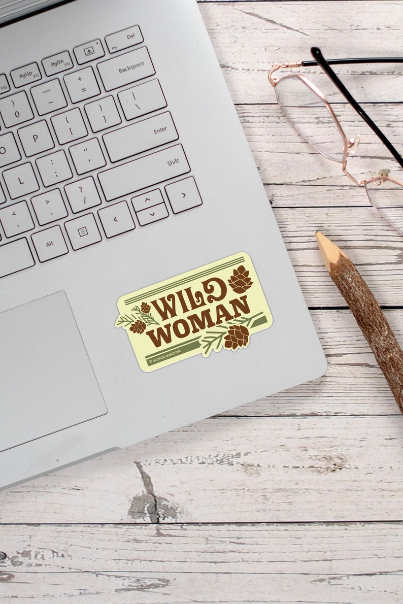 May include: A rectangular sticker on a laptop with the words "WILD WOMAN" in brown, surrounded by pine cones and branches. The sticker has a light yellow background and a green border. A pencil and glasses are also visible.