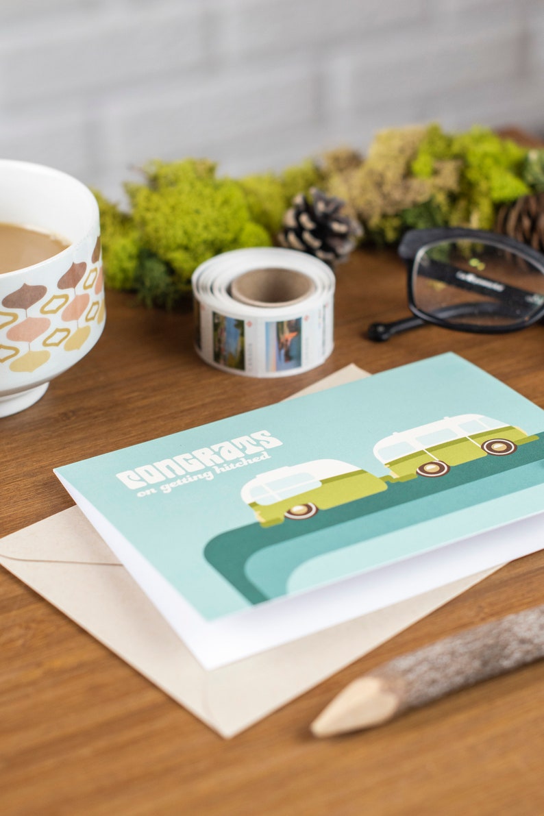 May include: A light blue card with the text "Congrats on getting hitched" and a green and white illustration of two camper vans. The card is sitting on a brown envelope.