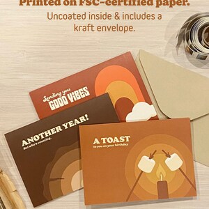 May include: Four blank greeting cards with different designs. The cards feature a rainbow, a campfire, and the text "Sending you good vibes", "Another year! But who's counting", and "A toast to you on your birthday".