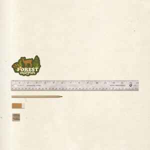 May include: A green and brown sticker with a deer and trees, and the text "FOREST explorer". A silver ruler with black markings, a wooden pencil, and two pencil sharpeners are on a white background.