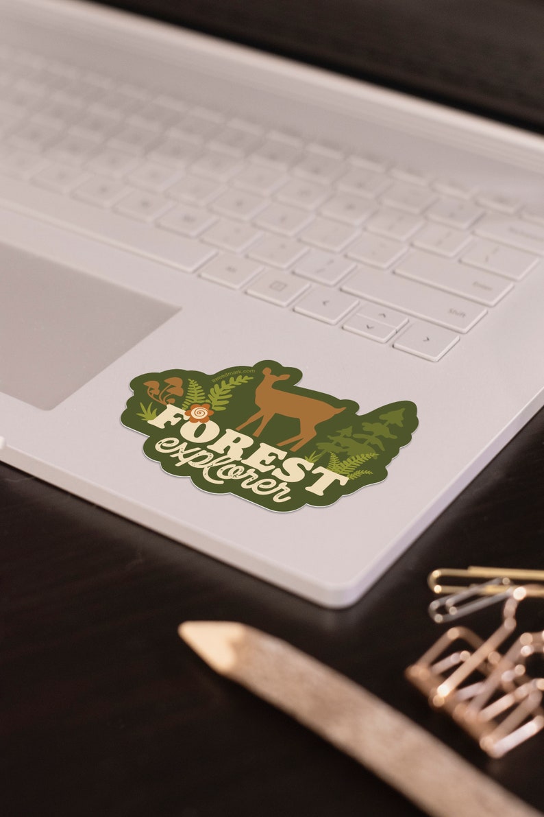 May include: A green and brown sticker with a deer silhouette and the text "FOREST explorer" in a retro font. The sticker is on a white laptop.