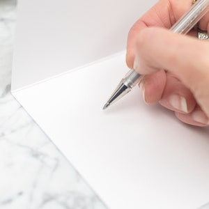 May include: A close-up of a blank white card with a silver pen writing on it. The card is on a white marble surface.