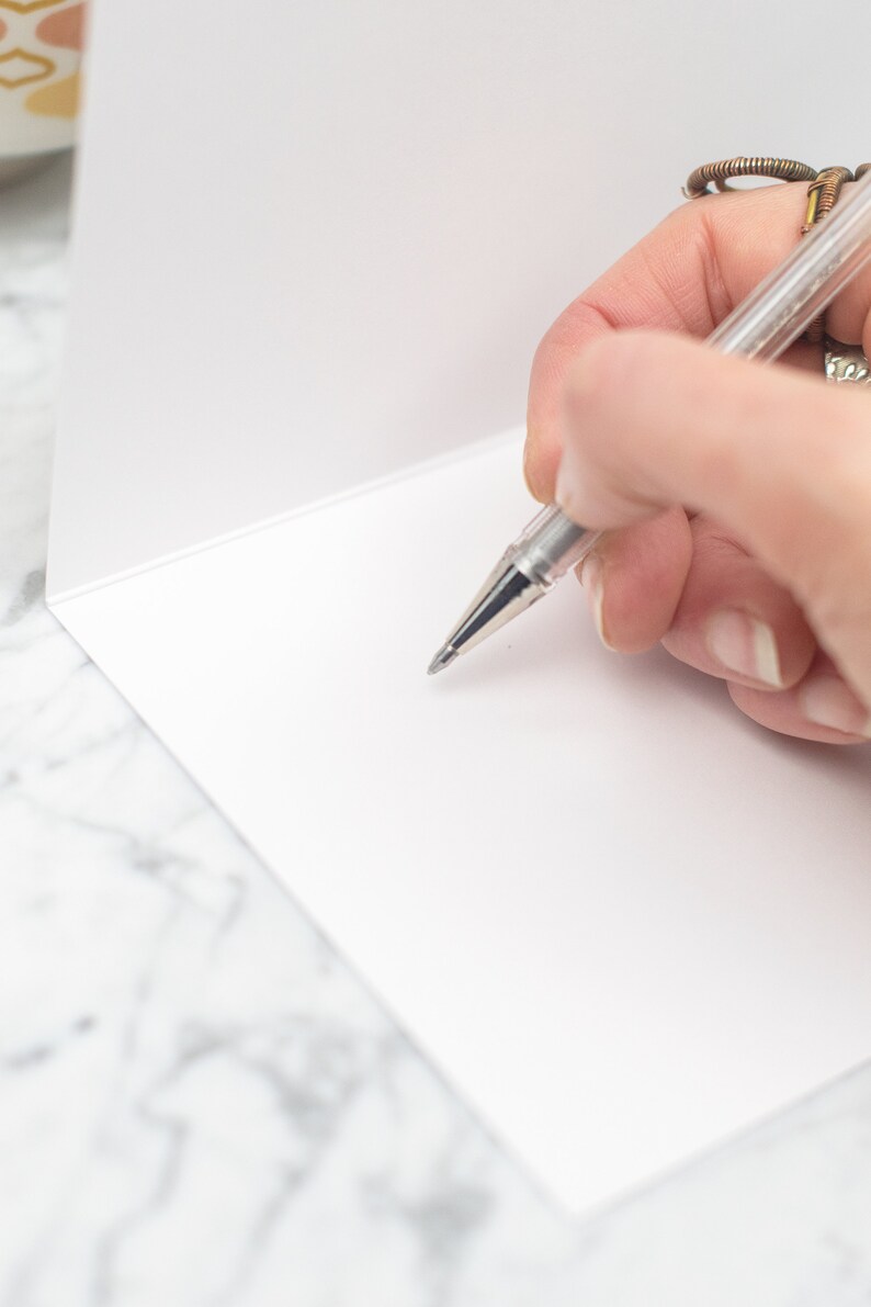 May include: A close-up of a blank white card with a silver pen writing on it. The card is on a white marble surface.