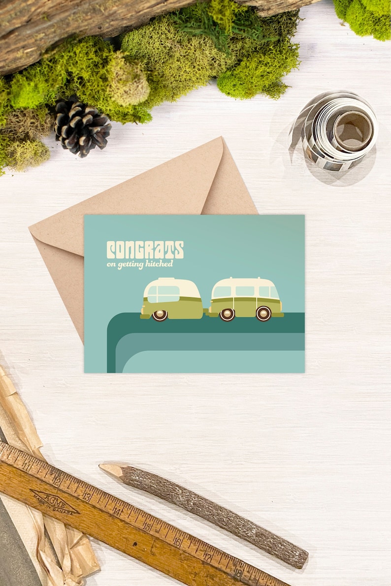 May include: A light blue card with the text "Congrats on getting hitched" and a cartoon illustration of two green and white camper vans. The vans are driving on a blue and green road.