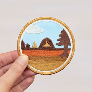 May include: A round embroidered patch depicting a campsite scene in the woods. The patch features a brown tent, a campfire, a tree, and a cloud in a blue sky. The patch is surrounded by a gold border and a white inner border.