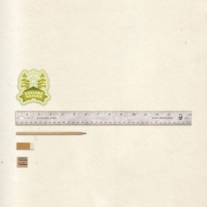 May include: A white ruler with black markings, a wooden pencil, and two pencil sharpeners on a beige background. The ruler is labeled "Stainless Steel" and "Acier Inoxydable". The ruler is 15 inches long.