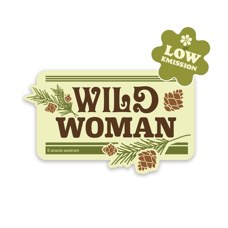 May include: A sticker with the words "WILD WOMAN" in brown, stylized font, with pine cones and pine needles. The sticker has a light yellow background and a green border. A green flower-shaped emblem reads "LOW EMISSION".
