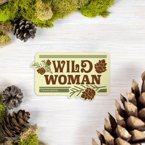 May include: A rectangular sticker with the words "WILD WOMAN" in brown, adorned with pine cones and branches. The sticker is set against a backdrop of moss, pine cones, and wood, creating a natural, outdoorsy theme.