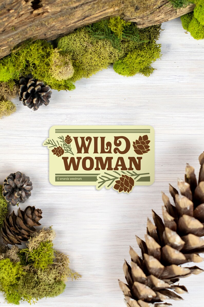 May include: A rectangular sticker with the words "WILD WOMAN" in brown, adorned with pine cones and branches. The sticker is set against a backdrop of moss, pine cones, and wood, creating a natural, outdoorsy theme.