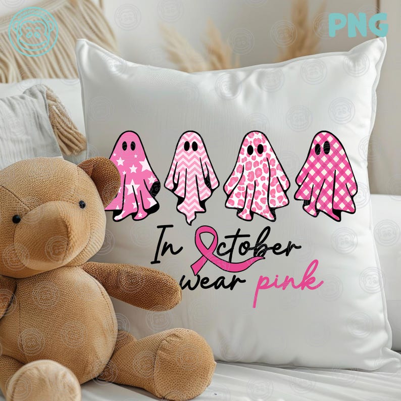 Op de afbeelding: Wit kussen met vier roze spookillustraties met verschillende patronen. Het kussen heeft de tekst "In October wear pink" met een roze lint. Een teddybeer staat op de voorgrond.