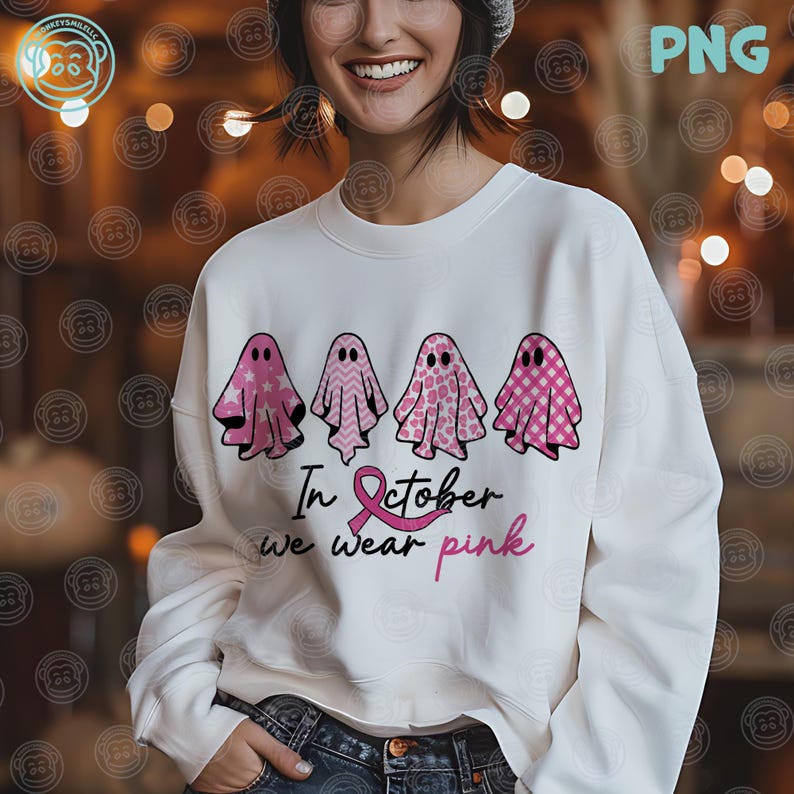 Op de afbeelding: Witte sweatshirt met vier roze spookillustraties met verschillende patronen. De tekst "In October we wear pink" staat eronder, samen met een roze lint. Het ontwerp is voor borstkankerbewustzijn.