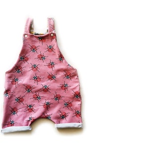 May include: Pink baby overalls with a ladybug print. The overalls have adjustable straps and a cuffed leg.