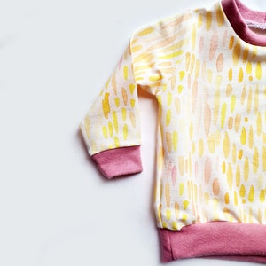 May include: A long-sleeved baby shirt with a white background and a yellow and orange watercolor pattern. The shirt has pink ribbed cuffs and a pink ribbed hem.