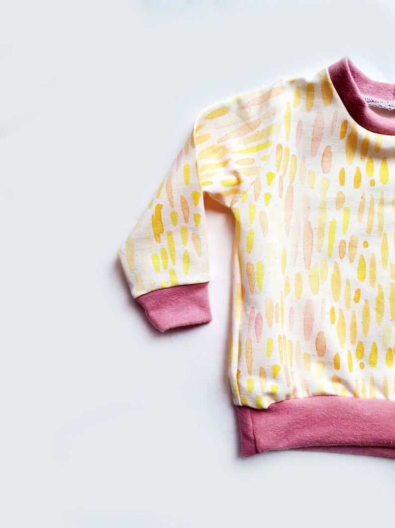 May include: A long-sleeved baby shirt with a white background and a yellow and orange watercolor pattern. The shirt has pink ribbed cuffs and a pink ribbed hem.