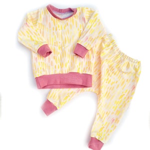 May include: A yellow and pink baby pajama set with a watercolor-like pattern of dots and dashes. The set includes a long-sleeved shirt and matching pants.