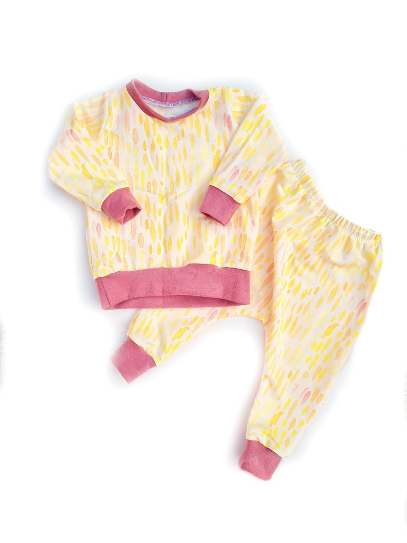 May include: A yellow and pink baby pajama set with a watercolor-like pattern of dots and dashes. The set includes a long-sleeved shirt and matching pants.